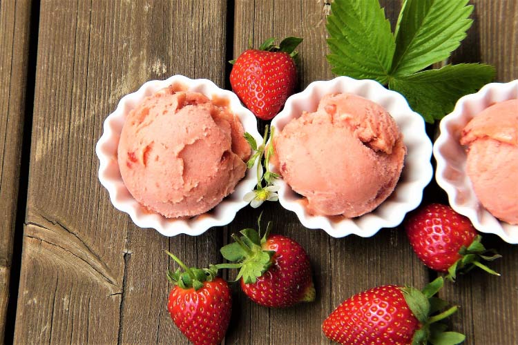 Aerial view of 3 white bowls of strawberry ice cream by fresh strawberries and mint leaves