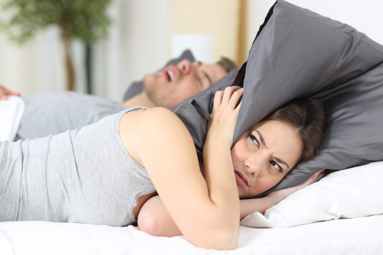Brunette woman covers her ears with her pillow in frustration due to her snoring husband with sleep apnea