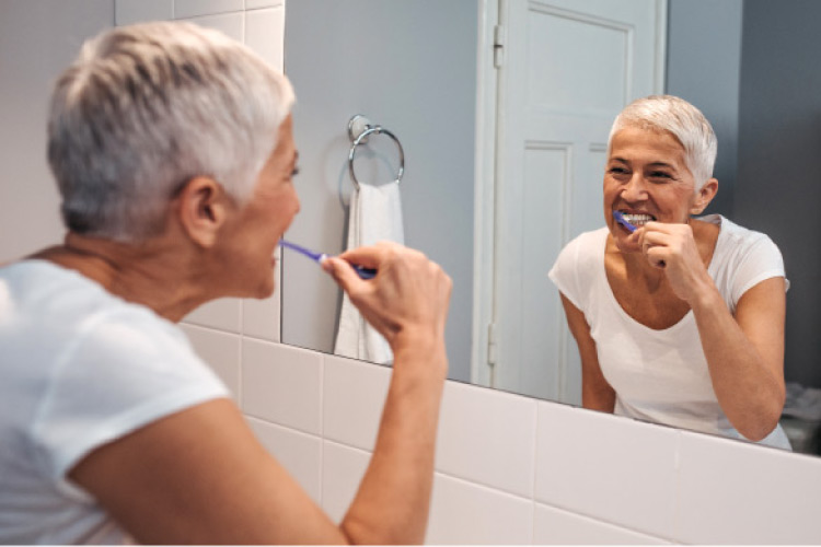 older woman brushing her teeth in a mirror to preserve her restorative dentistry