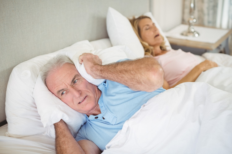 Older couple in bed with the woman snoring away and the man wide awake and covering his ears with a pillow