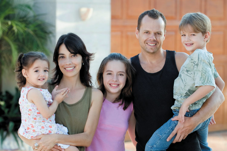 A smiling family standing in front of a home includes the mother holding a toddler girl, a father holding a young boy and young teen girl standing between them.