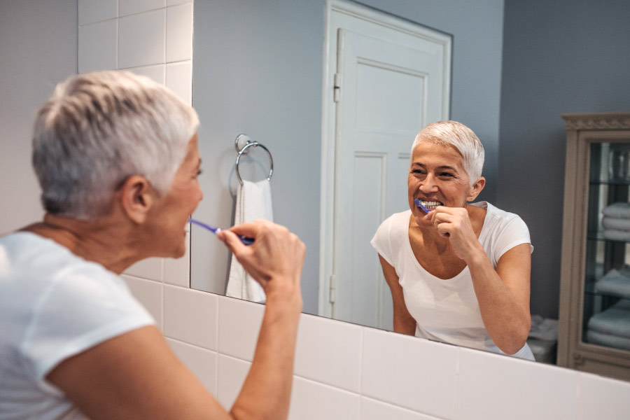 Lady with short gray hair brushing her teeth while looking in the mirror 