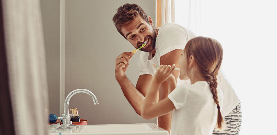 Side view of a dad & young daughter standing in front of the sink brushing their teeth